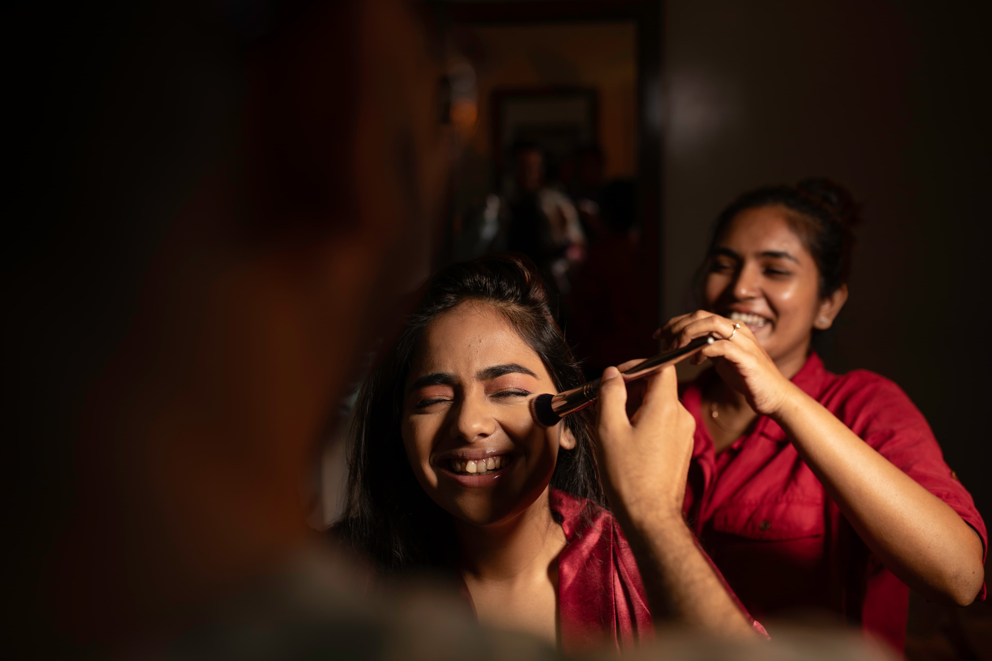 A bride laughing as her makeup is applied backstage by Couple Artistry by Shaash